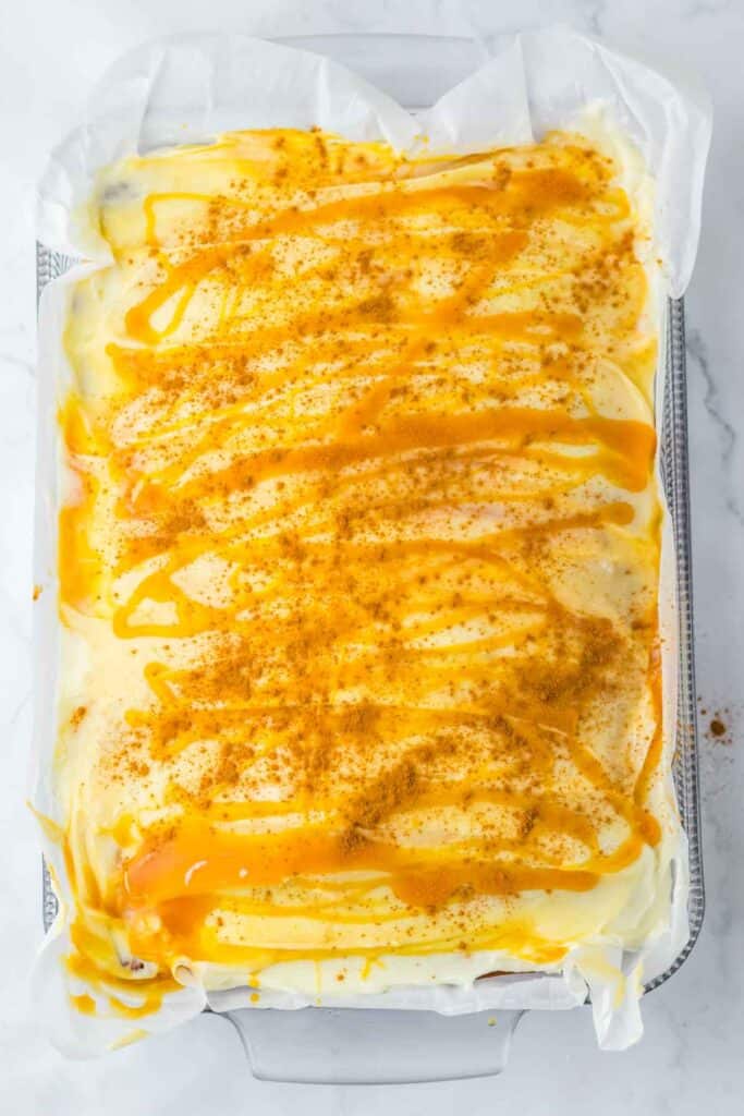 A rectangular baking dish filled with a creamy Pumpkin Caramel Cake, topped with a drizzle of caramel sauce and sprinkled with brown powder, likely cinnamon or cocoa. The dessert is lined with parchment paper.