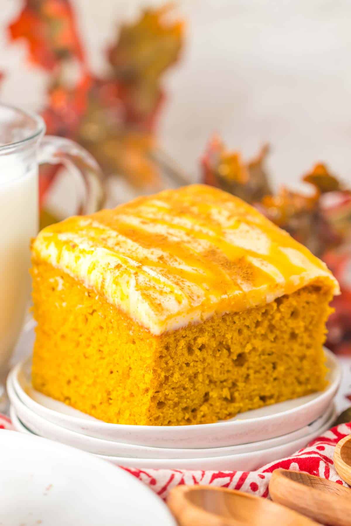 A slice of Pumpkin Caramel Cake topped with frosting and drizzle, served on a white plate, with autumn leaves blurred in the background.