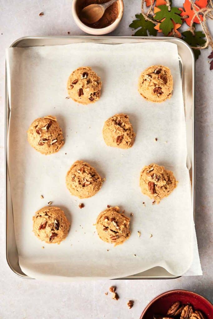 A baking tray with eight unbaked pumpkin cookies with pecans topped with chopped nuts on parchment paper. A small bowl with a spice mix and leaves are visible in the background.