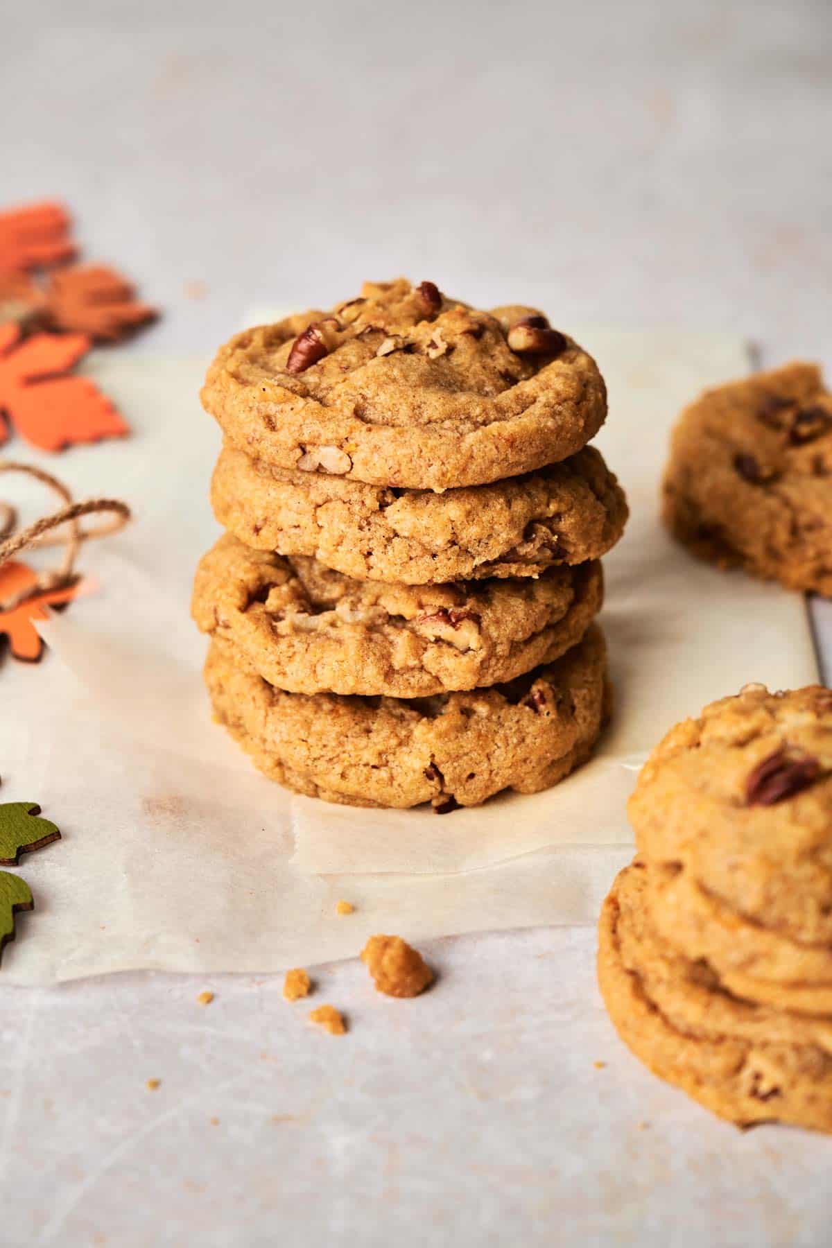 A stack of five Pumpkin Cookies With Pecans sits on parchment paper. More cookies are placed around, and autumn leaves are in the background.
