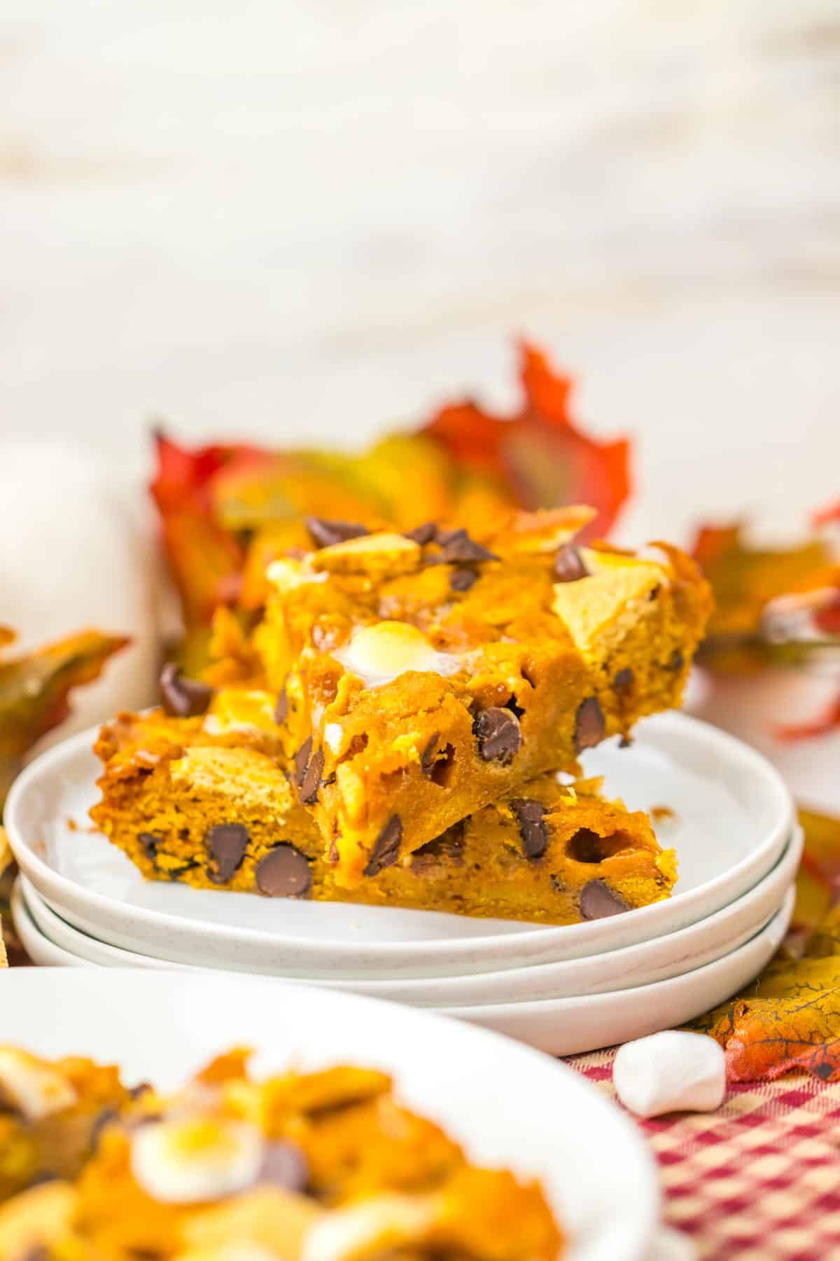 A stack of pumpkin s'mores cookie cake on a white plate, with autumn leaves in the background evokes the cozy flavors of a Pumpkin S’mores Cookie Cake.