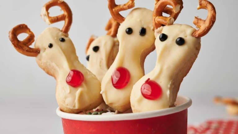 Cookies decorated to look like reindeer with pretzel antlers, black eyes, and red noses are placed in a red container.