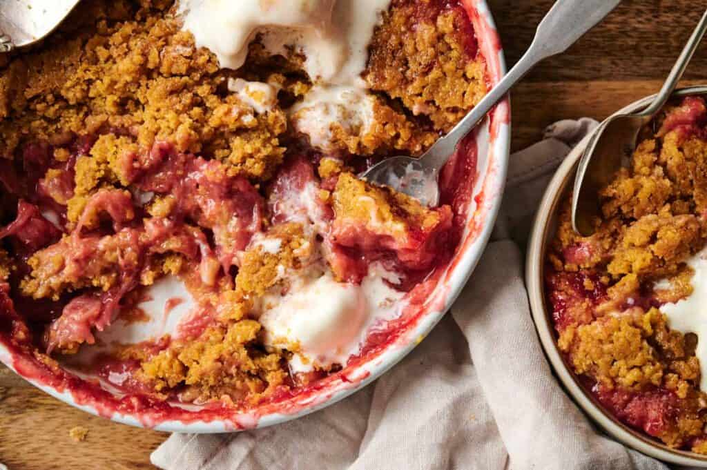 A baked fruit crumble with a golden-brown topping sits beside a Chocolate Pumpkin Patch Brownie Cake, both served with vanilla ice cream in bowls on a wooden table with a cloth napkin and spoons.