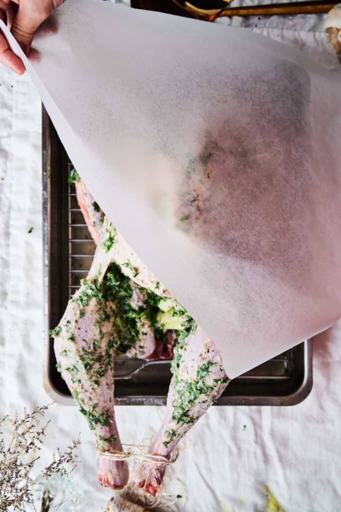 A person places parchment paper over a raw, marinated turkey in a roasting pan. The roasted turkey is coated with herbs and seasonings.