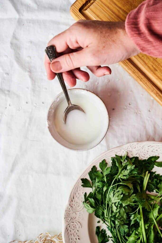 A hand holding a spoon over a small bowl of white liquid on a table with a plate of fresh parsley, a wooden cutting board, and slices of roasted turkey.