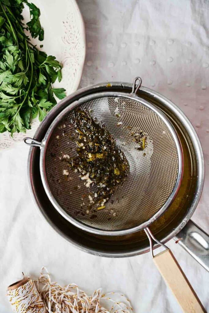 A metal strainer holding drained herbs and garlic rests atop a pot, with fresh parsley on a plate and twine with a spool nearby on a white surface, perfect ingredients for preparing roasted turkey.