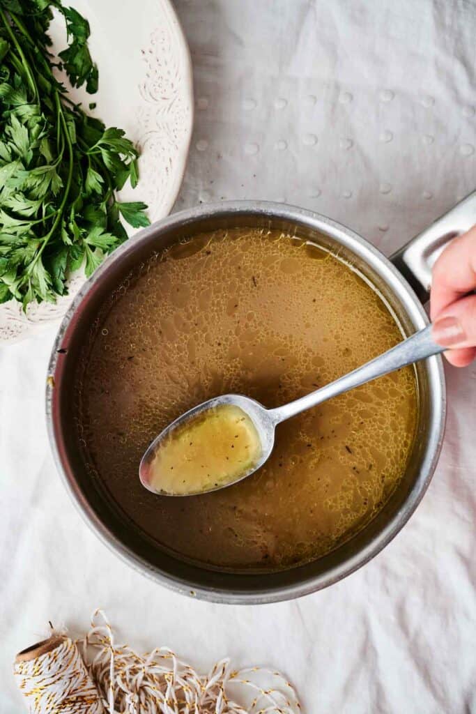 A hand holds a spoon over a pot of brown broth on a white surface, with a bunch of fresh parsley next to the pot, evoking the rich aroma of roasted turkey.