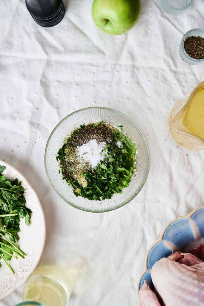A bowl filled with chopped herbs, ground pepper, and salt sits on a table alongside an apple, some parsley, a dish containing raw meat for roasted turkey, and a large glass of sauce.