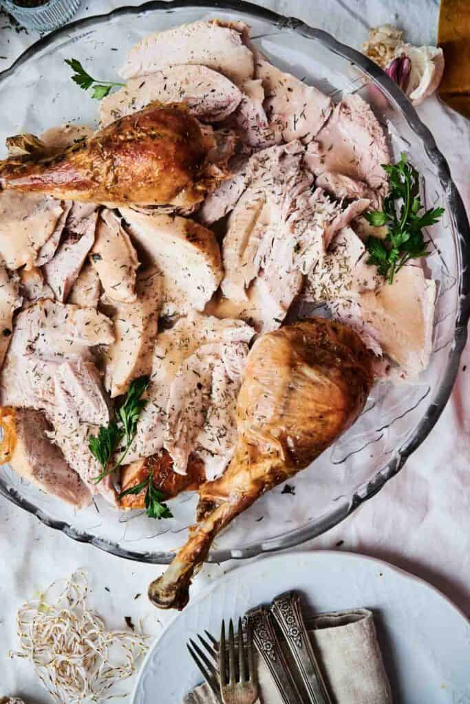 A platter of succulent roasted turkey slices, including two delectable drumsticks, garnished with sprigs of parsley. Silverware and a napkin are arranged beside the plate on a pristine white tablecloth.