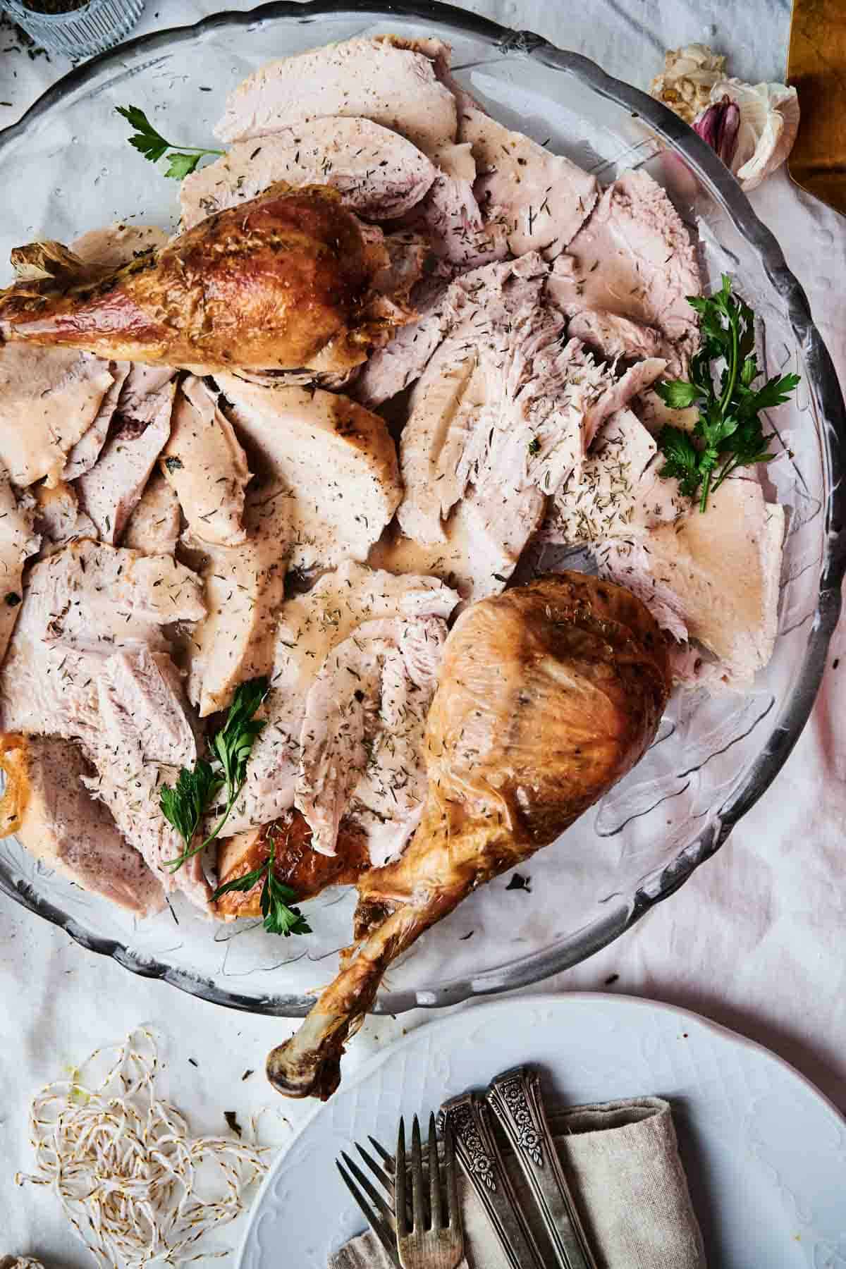 A platter of succulent roasted turkey slices, including two delectable drumsticks, garnished with sprigs of parsley. Silverware and a napkin are arranged beside the plate on a pristine white tablecloth.