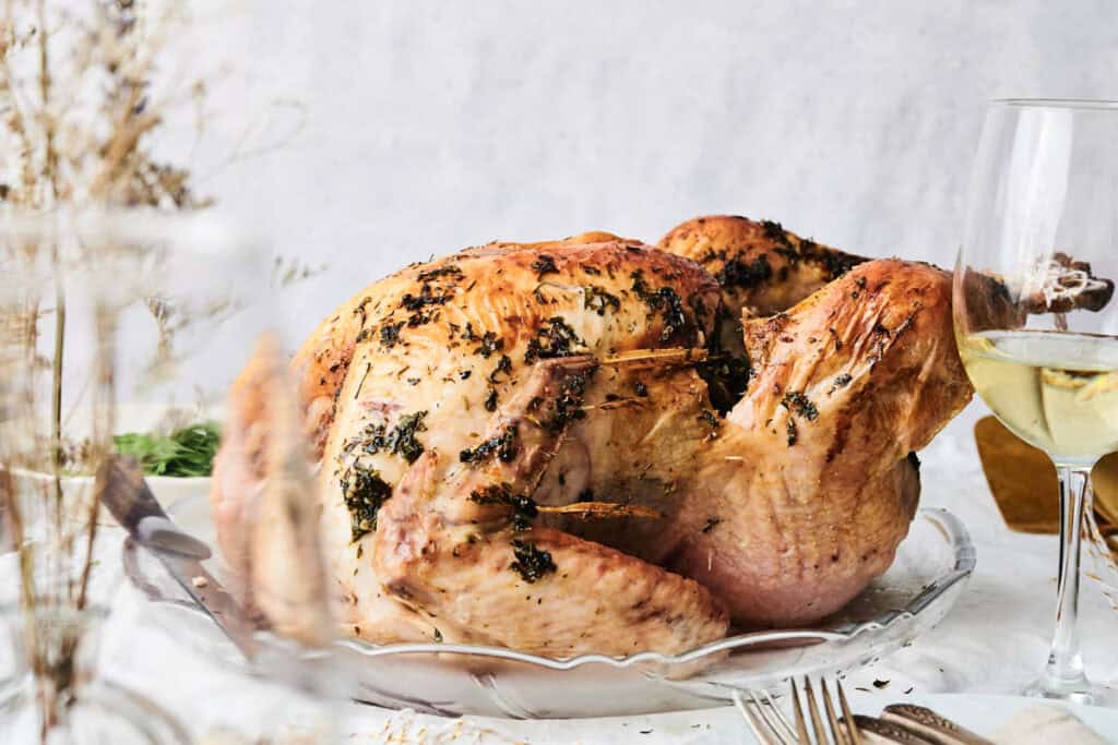 A succulent roasted turkey garnished with fresh herbs is artfully arranged on a glass platter, complemented by a glass of white wine.