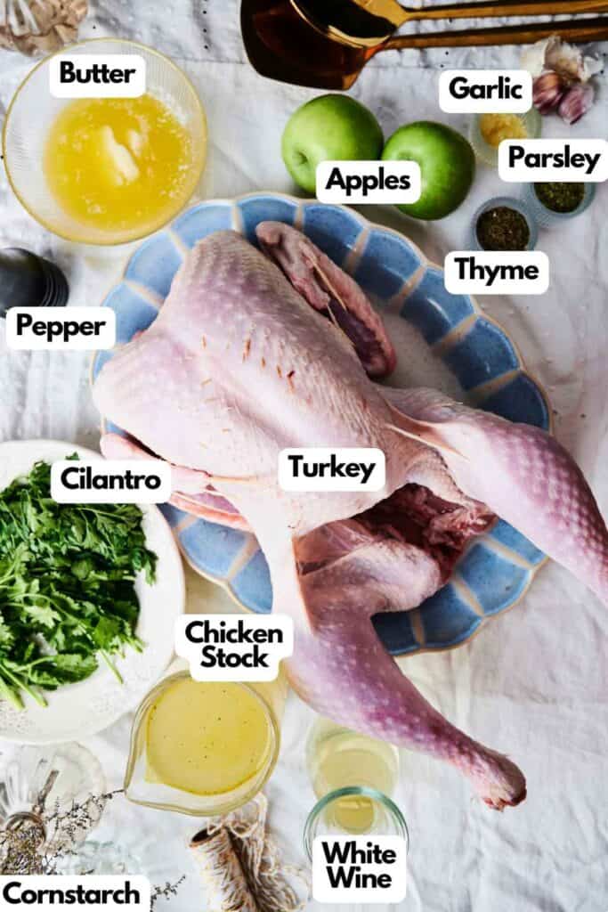 Overhead view of a raw turkey on a plate surrounded by various ingredients labeled: butter, apples, garlic, parsley, thyme, pepper, cilantro, chicken stock, cornstarch, and white wine&mdash;everything you need for a perfectly roasted turkey.