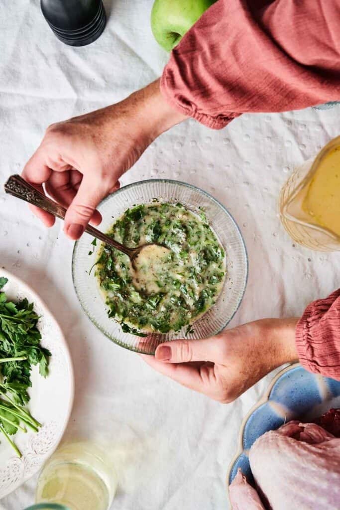 A person mixes a herb and mustard sauce in a bowl, placed on a table with fresh parsley, an apple, a pepper grinder, and raw poultry ready to become roasted turkey.