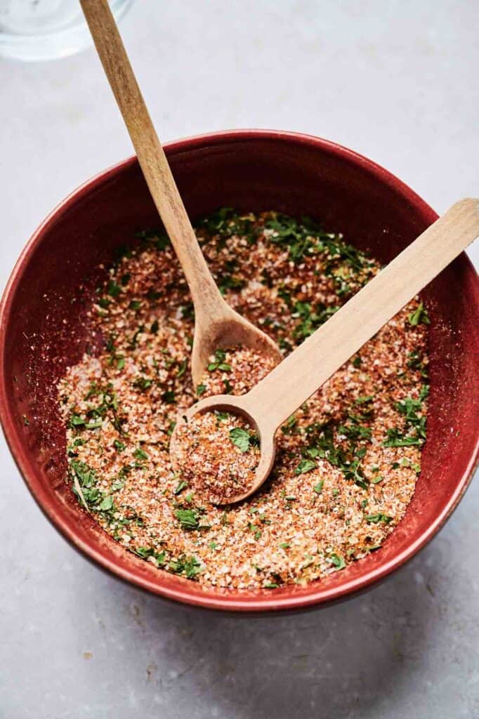 A red bowl filled with a dry herb and spice mixture reminiscent of Wingstop's Louisiana Rub sits invitingly, with two wooden spoons placed in the bowl.