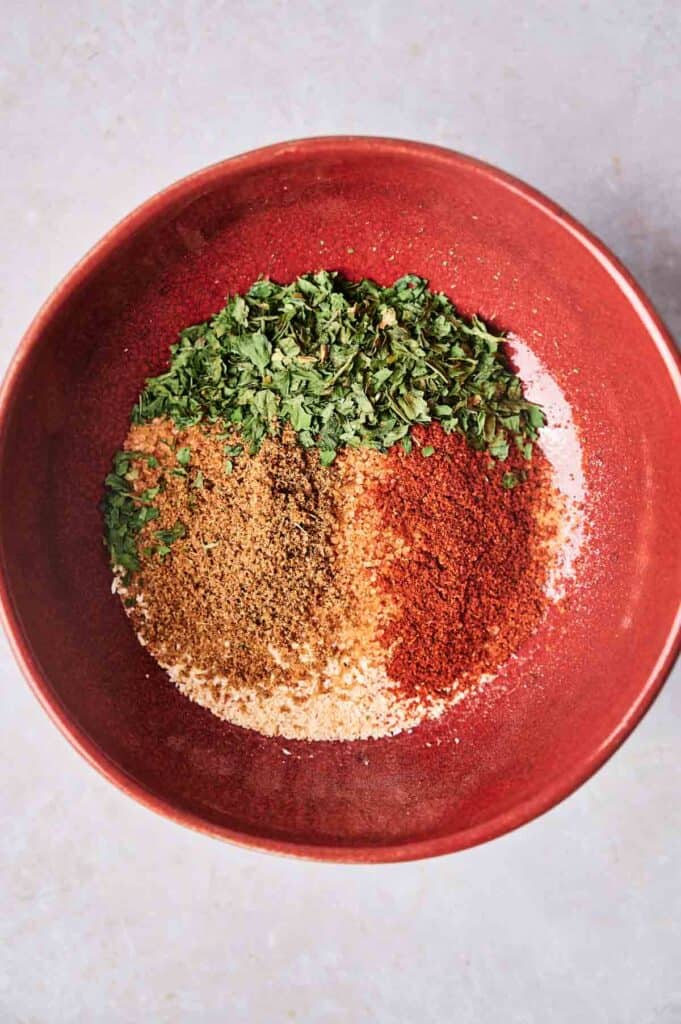 A red bowl containing a mixture of chopped herbs and ground spices, reminiscent of Wingstop's Louisiana Rub, with green herbs, brown spices, and red spices.