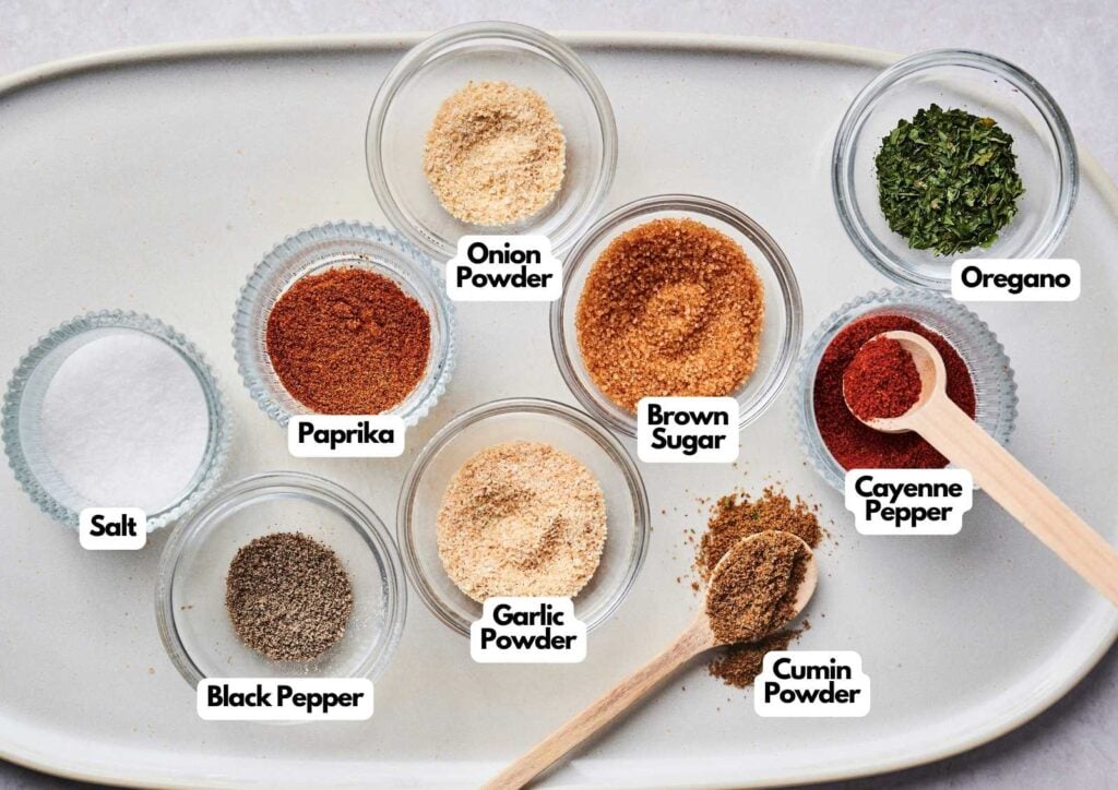 Overhead view of nine small glass bowls on a cream platter, each labeled with an ingredient for Wingstop Louisiana rub: oregano, cayenne pepper, cumin powder, brown sugar, onion powder, garlic powder, paprika, black pepper, and salt.
