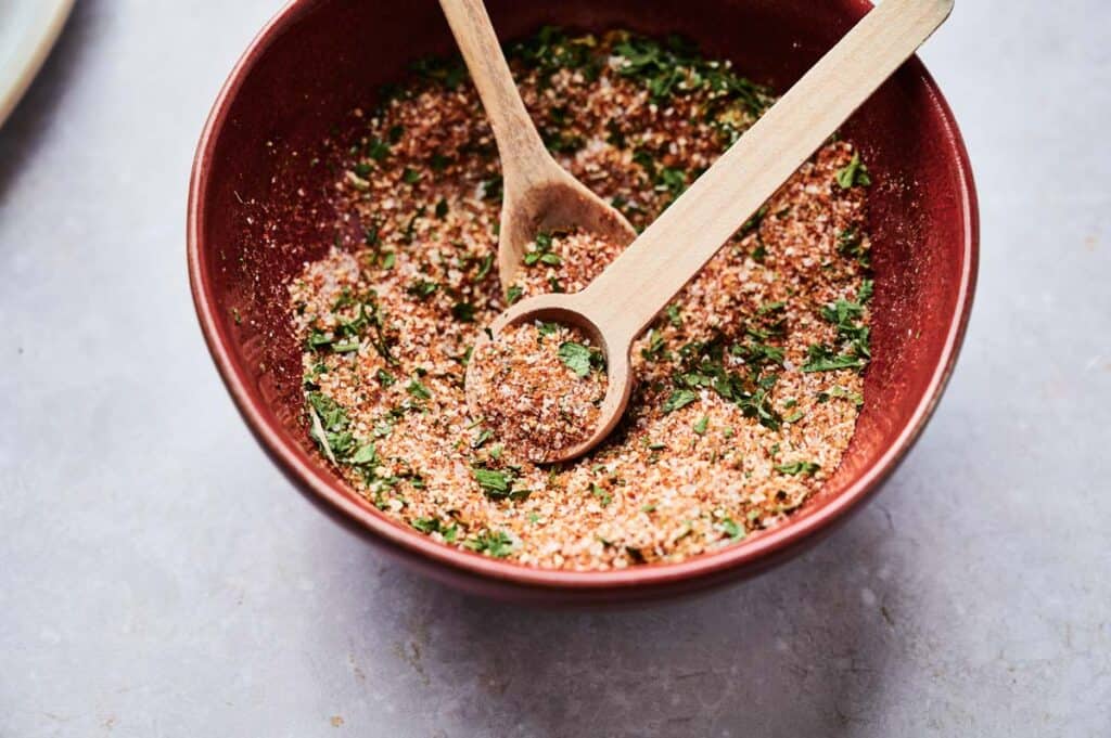 A ceramic bowl containing a dry spice mix with chopped herbs is shown with two wooden spoons resting inside, reminiscent of Wingstop Louisiana Rub.