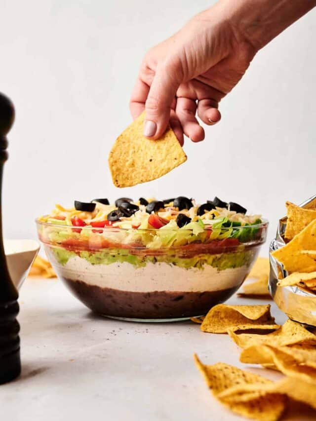 A hand dips a tortilla chip into a layered dip featuring beans, guacamole, sour cream, lettuce, tomatoes, cheese, and olives. A bowl of tortilla chips is nearby.
