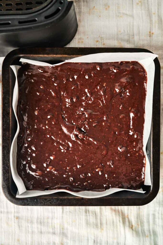 A square pan filled with unbaked chocolate brownie batter sits on a parchment-lined surface, ready for the magic of the air fryer to transform it into decadent Air Fryer Chocolate Brownies.