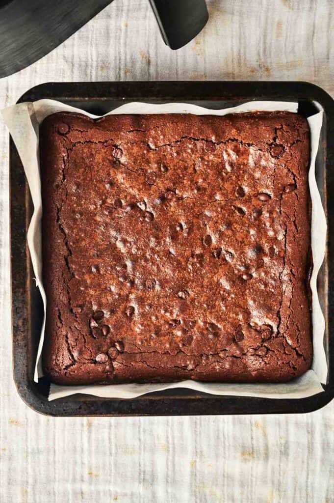 A square chocolate brownie with a slightly cracked surface rests in an air fryer baking pan lined with parchment paper.