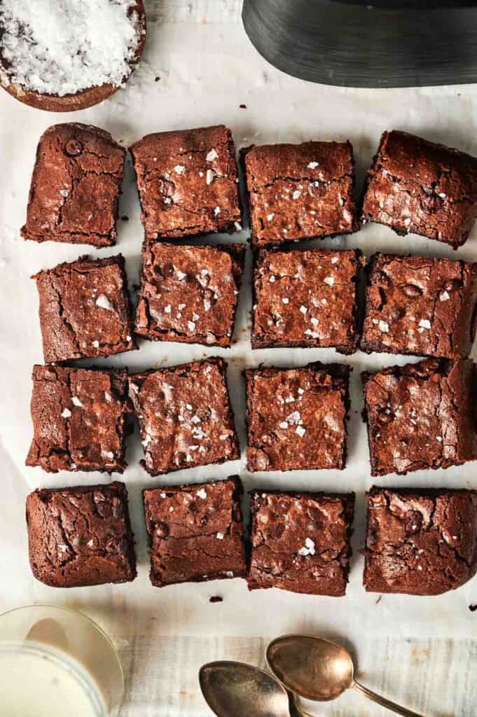 A batch of sixteen evenly cut air fryer chocolate brownies sprinkled with sea salt, placed on a parchment-lined surface. Nearby are a bowl of sea salt, a glass of milk, and three spoons.