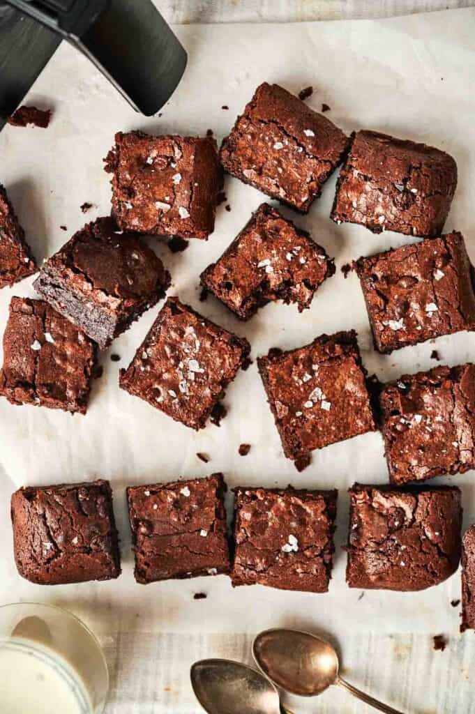 Squares of air fryer chocolate brownies with a sprinkle of sea salt rest on parchment paper, nestled beside a black container and two spoons.
