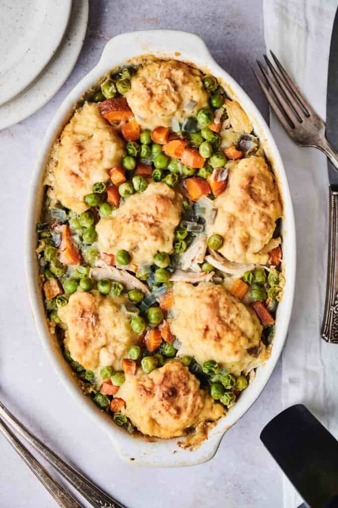 An air fryer chicken and dumpling casserole in an oval dish, topped with six biscuit-like dumplings, features peas, carrots, and shredded chicken. Forks and a napkin are beside the dish, with plates stacked nearby.