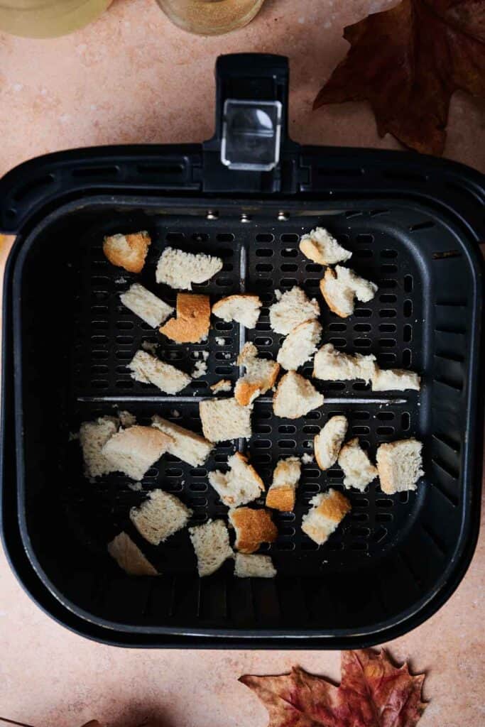 Cubed bread pieces nestled in an air fryer basket rest on a beige surface adorned with autumn leaves, evoking the cozy spirit of a Thanksgiving casserole in the making.