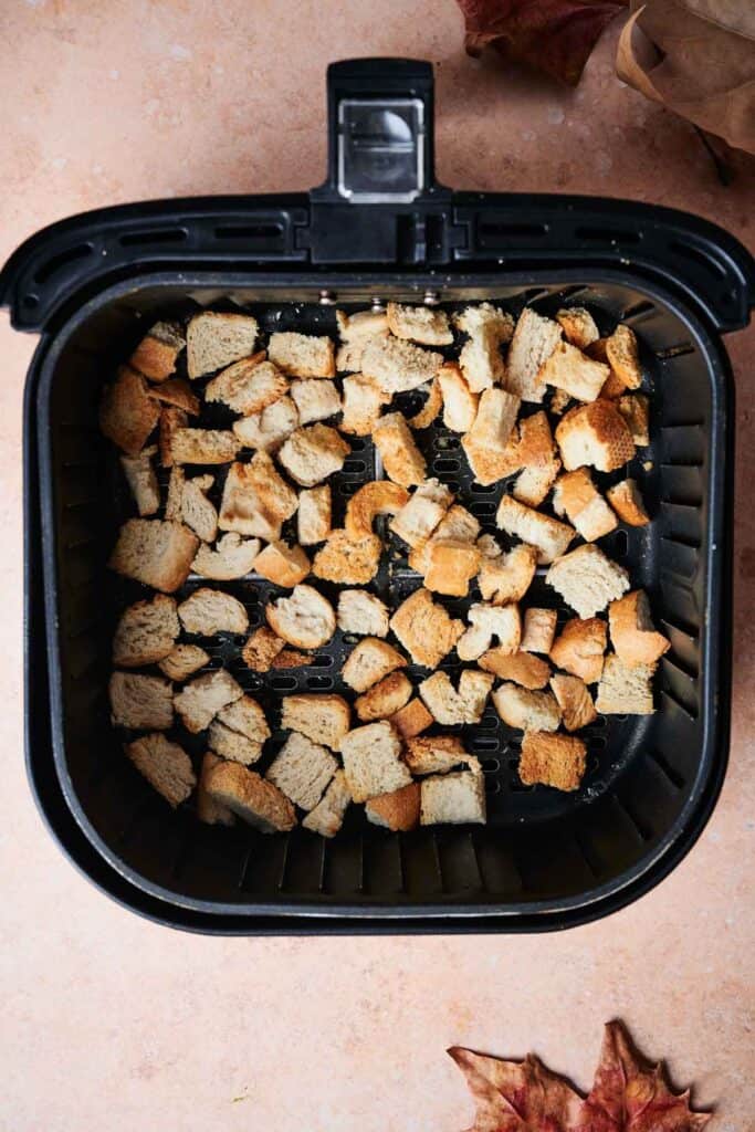 Crispy bread cubes in an air fryer basket rest on a light brown surface, surrounded by autumn leaves, ready to elevate your Thanksgiving casserole.
