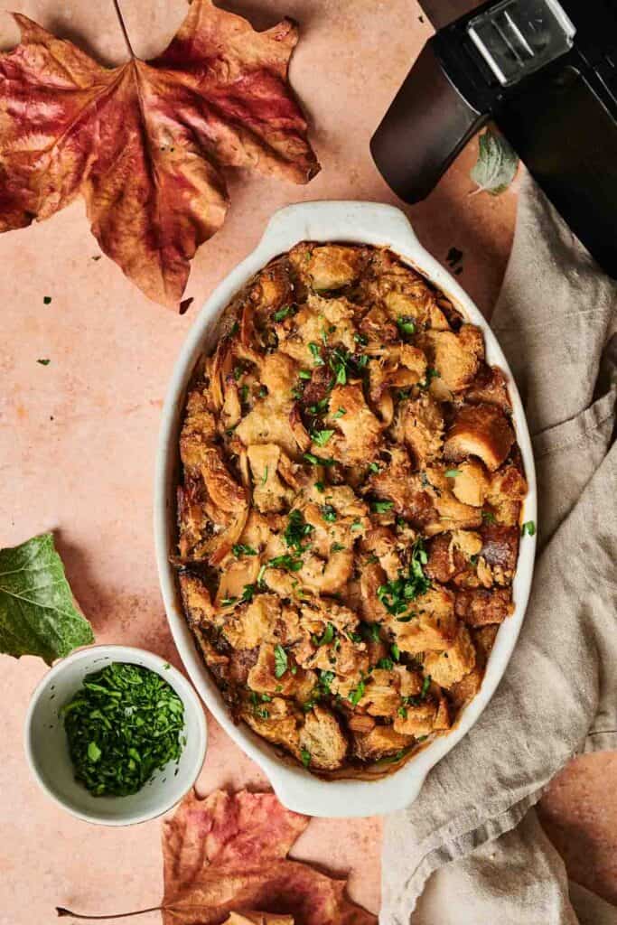 A dish of Thanksgiving casserole topped with chopped herbs, surrounded by autumn leaves and a small bowl of herbs, set on a beige surface. Perfectly made in an air fryer for that crispy finish.