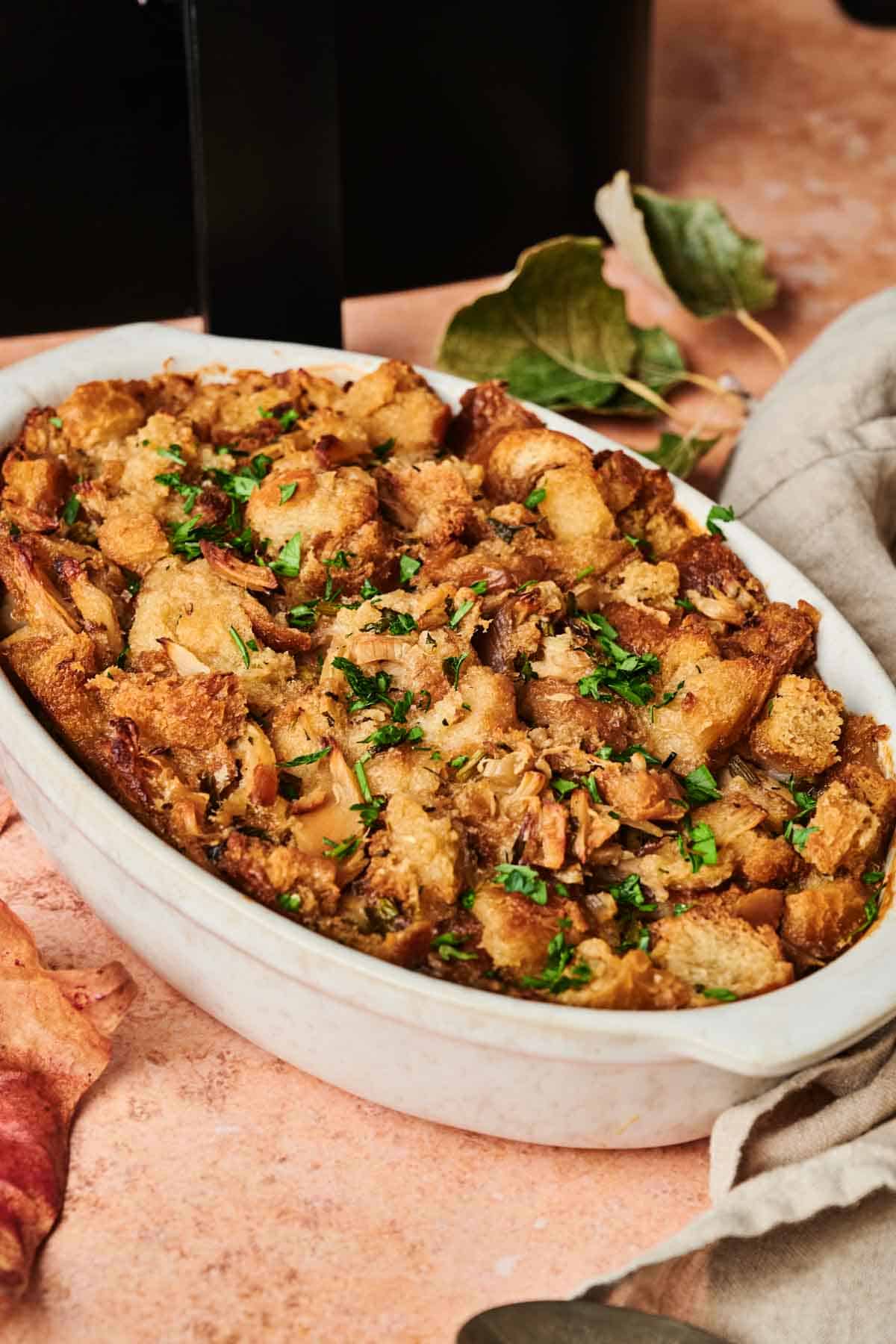 White dish filled with golden-brown stuffing, reminiscent of a classic Thanksgiving casserole, garnished with chopped parsley. It's set on a beige surface adorned with scattered autumn leaves, perfectly capturing the cozy holiday spirit. Consider using an air fryer for an extra crispy finish.