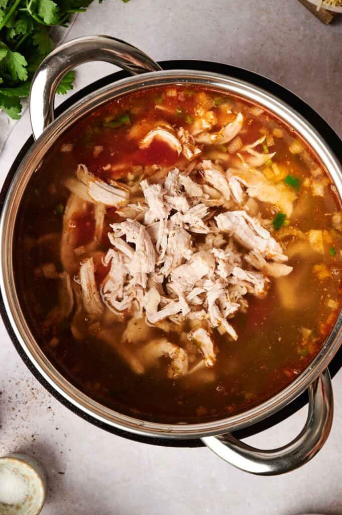 A pot filled with chicken tortilla soup, featuring shredded chicken, vegetables, and herbs, viewed from above on a gray surface.