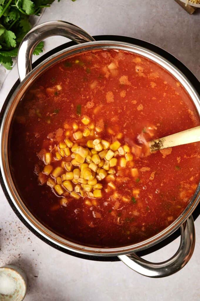 A pot of tomato soup with corn and herbs, reminiscent of a hearty chicken tortilla soup, being stirred with a wooden spoon.