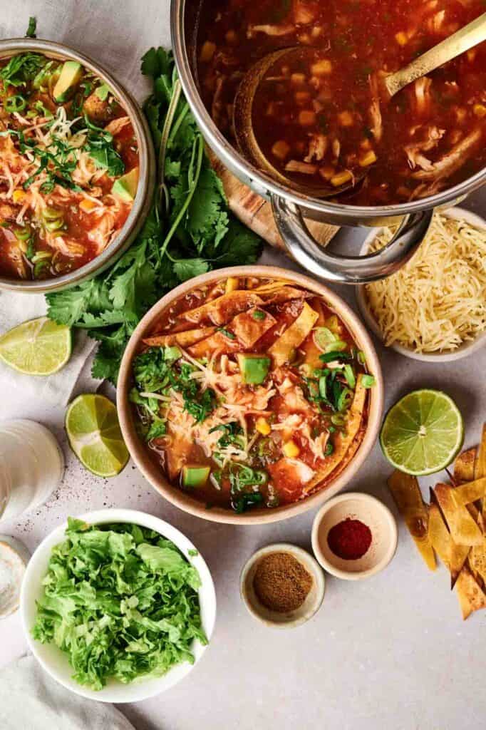 Bowls of chicken tortilla soup bursting with flavor are arranged on a table, featuring ingredients like lime, cilantro, shredded cheese, spices, and crispy tortilla strips.