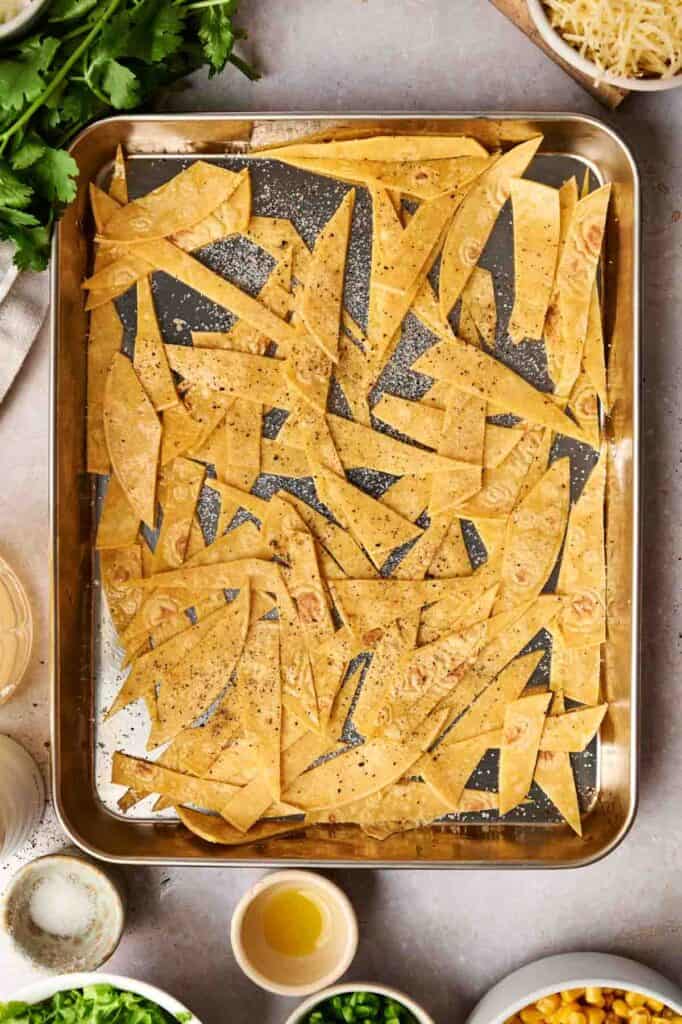 Tortilla strips seasoned with pepper are arranged on a baking tray, ready to complement a hearty bowl of chicken tortilla soup. Ingredients like cilantro, cheese, and green onions surround the tray, completing the perfect setting for a flavorful meal.