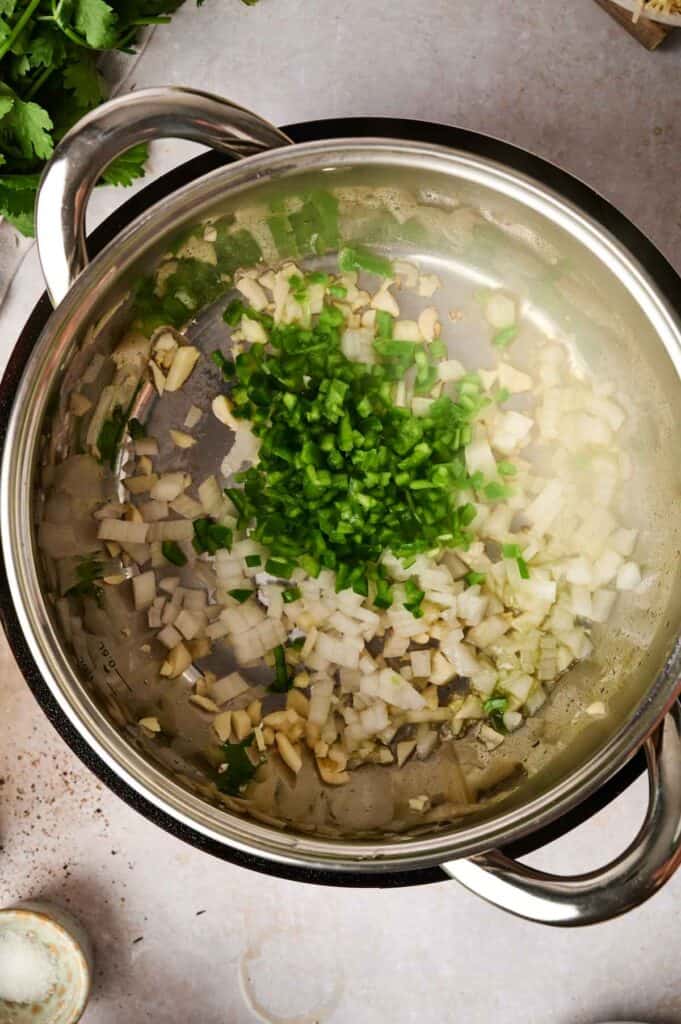 Chopped onions, garlic, and green peppers simmer in a stainless steel pot, creating the aromatic base for a hearty chicken tortilla soup.