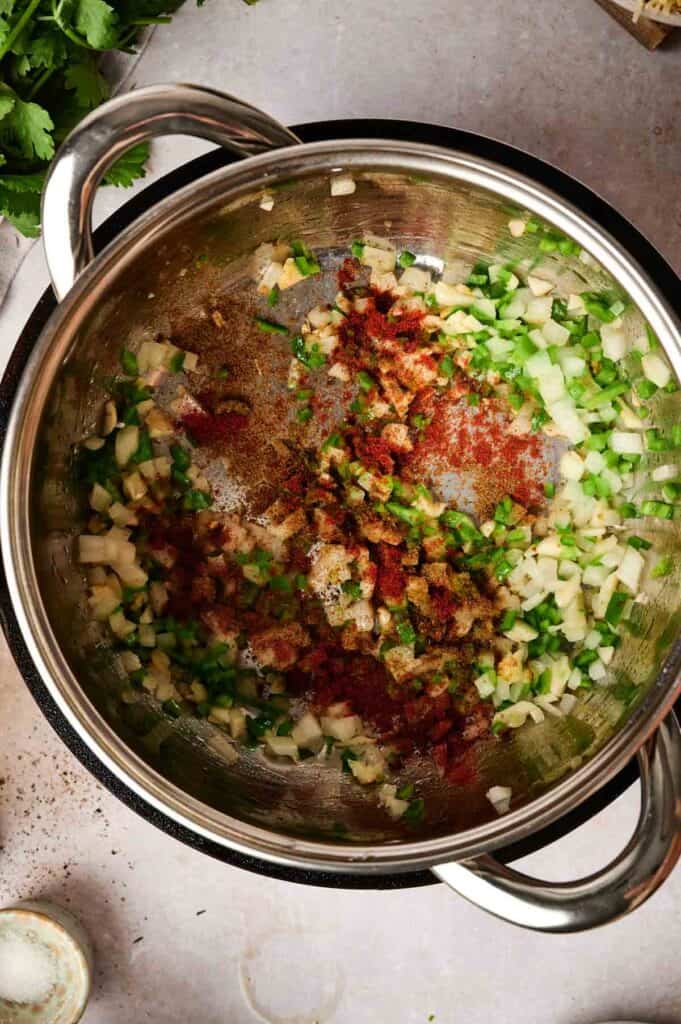 Chopped onions and green peppers with spices simmer in a pot of chicken tortilla soup on a stovetop, viewed from above.