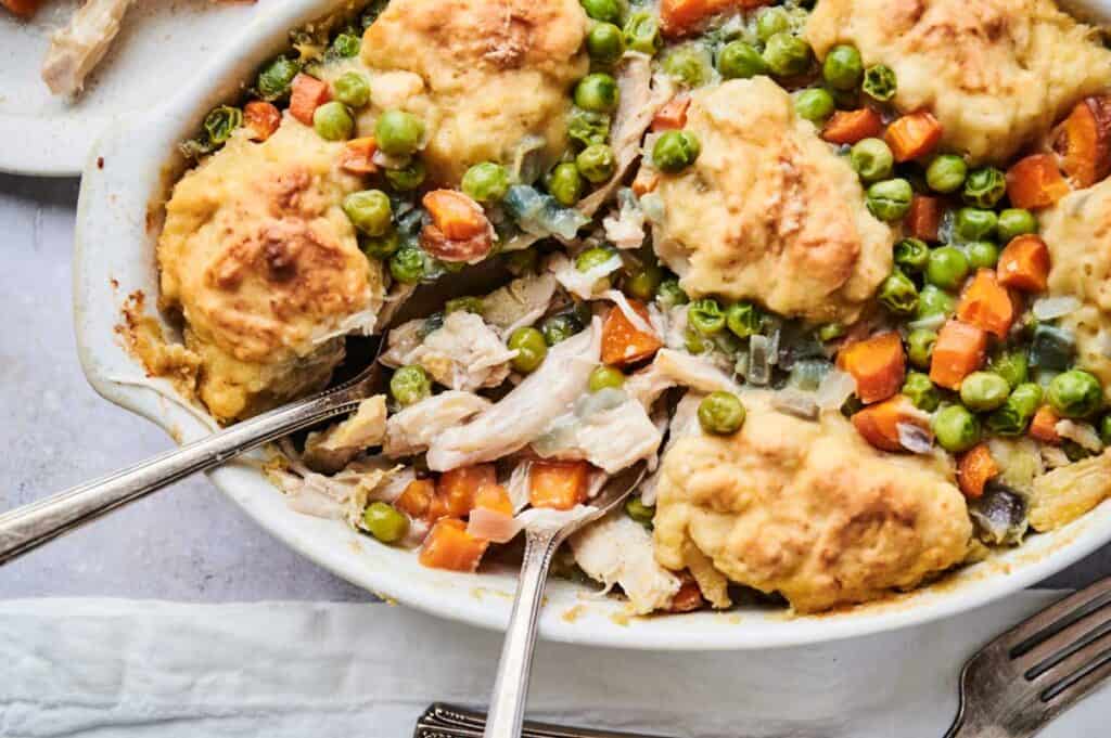 An Air Fryer Chicken and Dumpling Casserole featuring a savory blend of chicken, peas, and carrots is topped with golden brown biscuits. Two serving spoons rest in the dish, ready for a hearty helping.