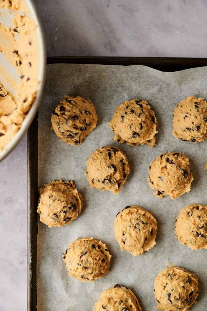 Cookie dough scoops with chocolate chips arranged on a parchment-lined baking sheet, next to a bowl of dough.