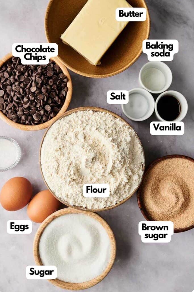 Ingredients for baking chocolate chip cookies arranged on a surface include butter, chocolate chips, baking soda, vanilla, salt, flour, brown sugar, eggs, and sugar.