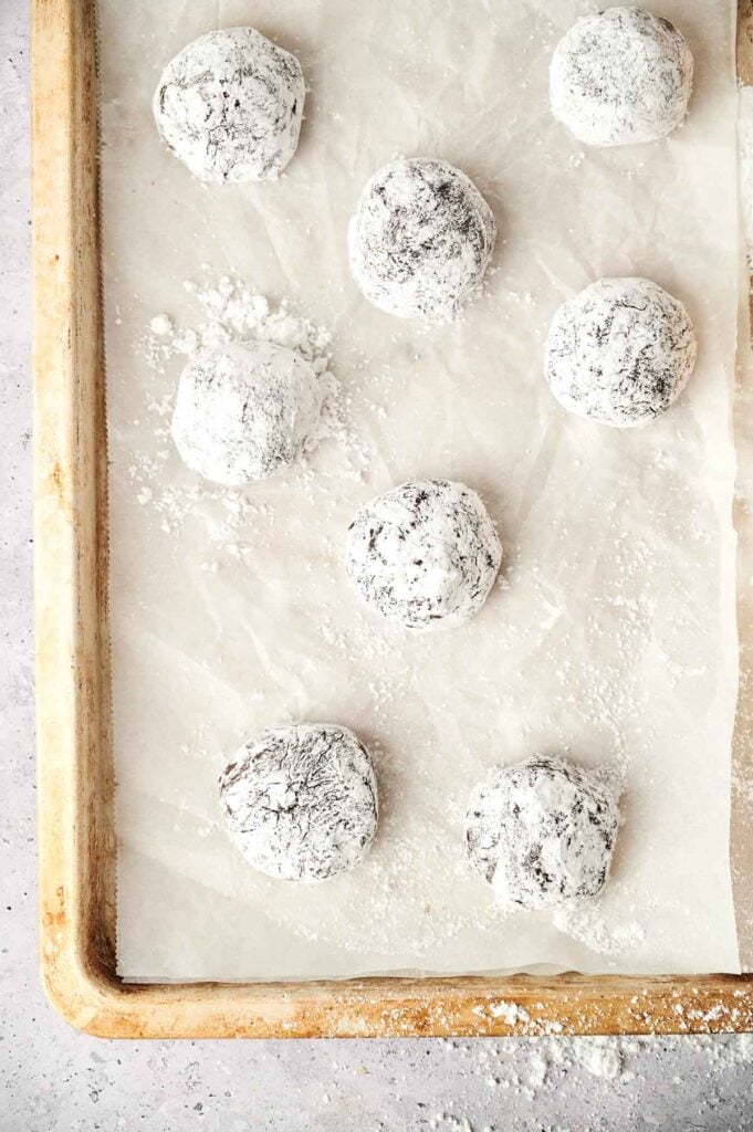 Nine unbaked crinkle cookies covered in powdered sugar on a parchment-lined baking sheet.