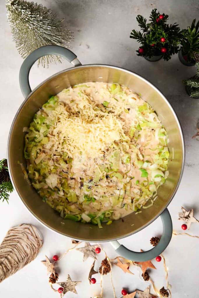 A large pot of Christmas casserole filled with chopped green vegetables and grated cheese, surrounded by decorative plants and holiday-themed items on a table.