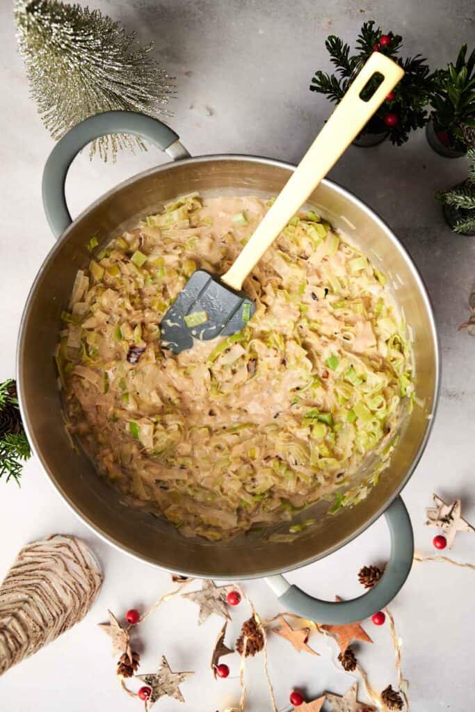 A pot of creamy pasta, transformed into a festive Christmas casserole, is mixed with chopped leeks and mushrooms. A spatula rests on the delicious mixture as holiday decorations surround the pot.