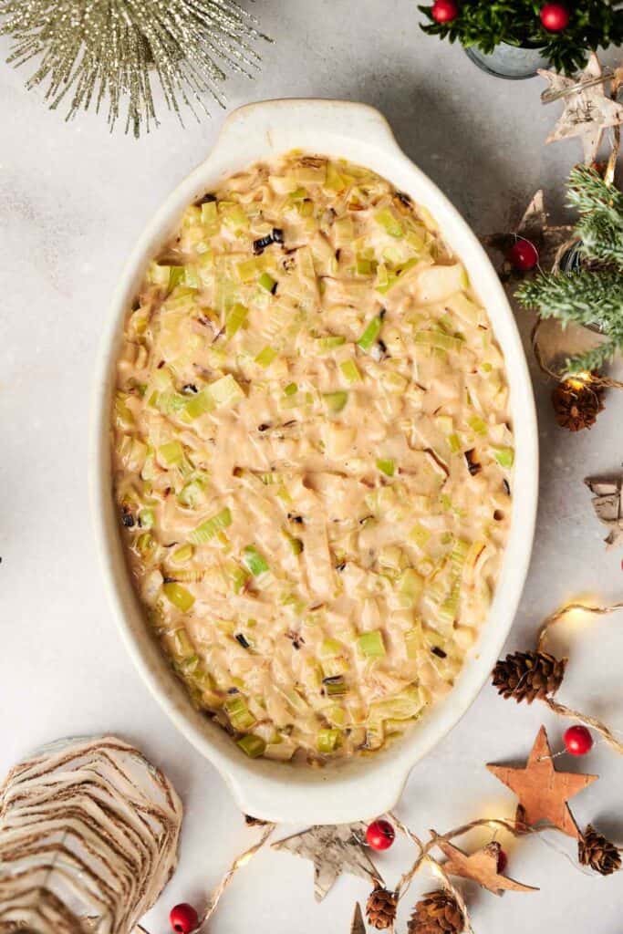 Oval dish featuring a Christmas casserole, with a baked creamy center topped with chopped green vegetables. It’s surrounded by festive decorations like pinecones and red berries, capturing the holiday spirit perfectly.