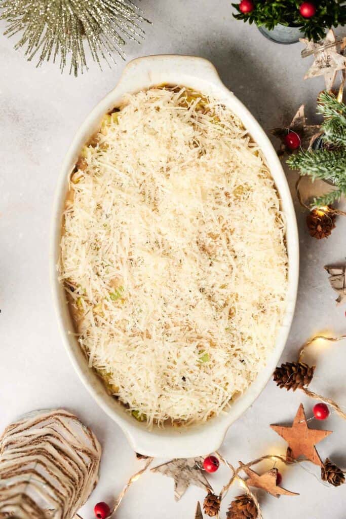 A Christmas Casserole sits in an oval dish with grated cheese on top, surrounded by festive holiday decorations like pinecones, red berries, and star ornaments on a light surface.