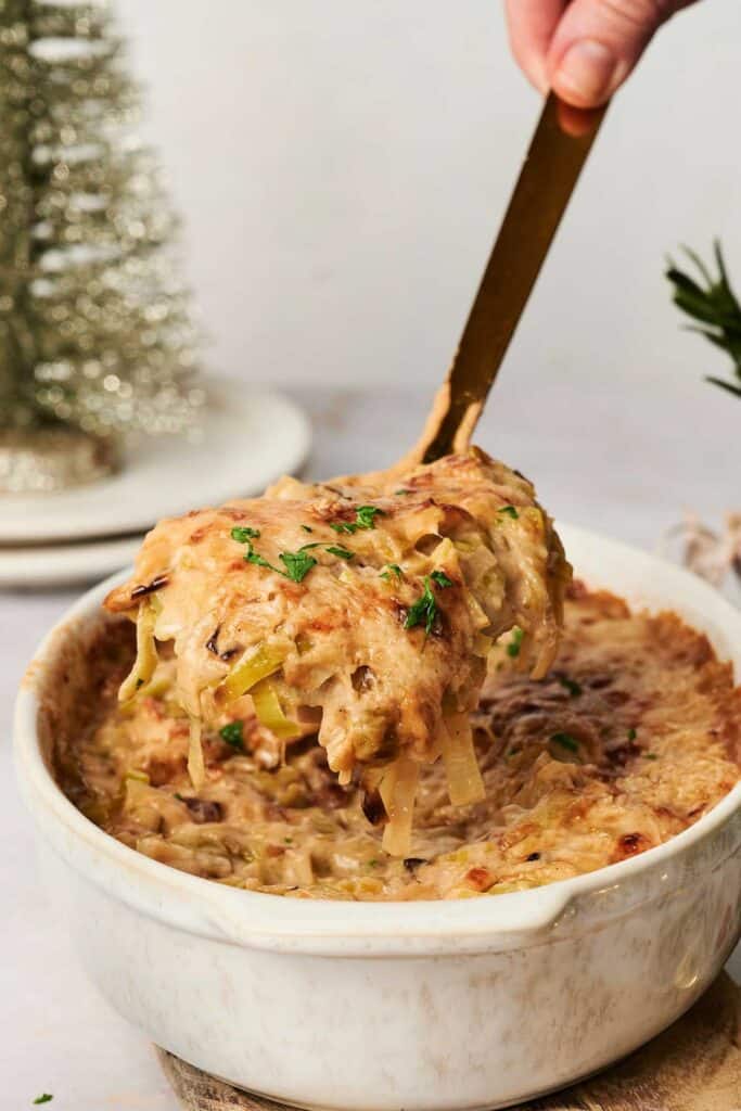 A hand serving a portion of baked creamy vegetable pasta from a white casserole dish, garnished with herbs, beside a festive Christmas decoration.