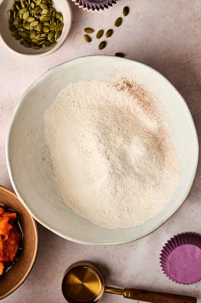 A bowl of flour and spices sits on a countertop surrounded by pepitas, pumpkin purée, a measuring cup, and empty cupcake liners—everything you need to create homemade treats inspired by Starbucks Pumpkin Cream Cheese Muffins.