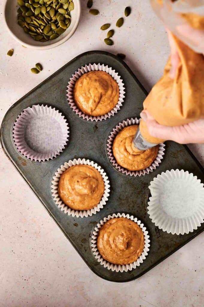 Piping pumpkin batter into paper-lined muffin tin cups on a speckled countertop, with a bowl of pumpkin seeds nearby.