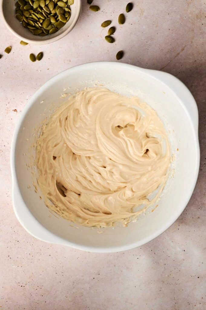 White mixing bowl with creamy batter on a light surface, with a small bowl of pumpkin seeds and scattered seeds nearby.