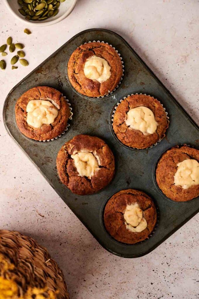 A muffin tray with six pumpkin muffins topped with cream cheese is on a countertop. A small bowl of pumpkin seeds is nearby.